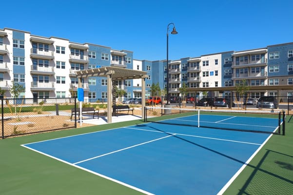 Tennis court with blue surface and benches near residential buildings.