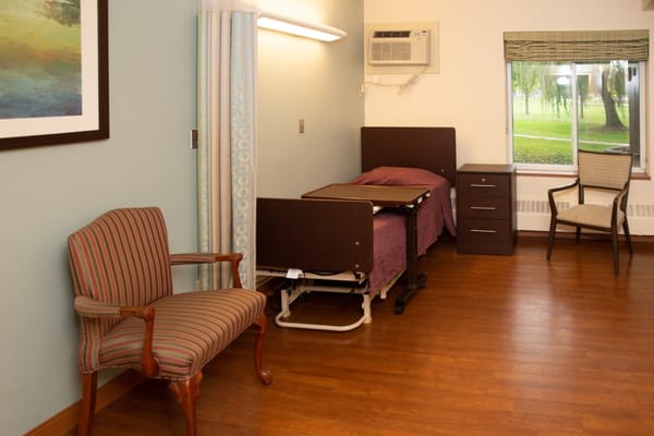 A cozy resident room featuring a bed, chair, and window.