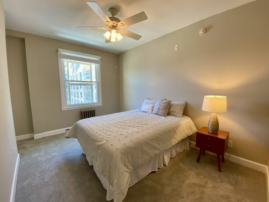 A well-lit bedroom featuring a bed with decorative pillows, a nightstand with a lamp, and a window view.
