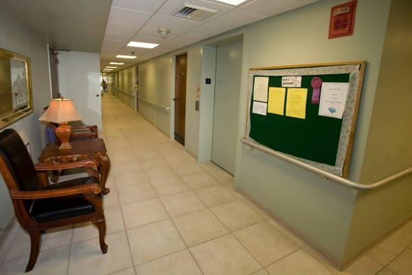 A well-lit hallway in Baptist Oaks Apartments featuring a chair, lamp, and bulletin board