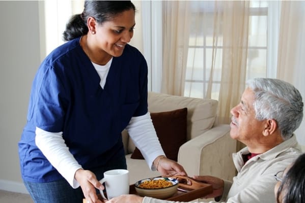 Caregiver handing a meal tray to an elderly man in a living room.