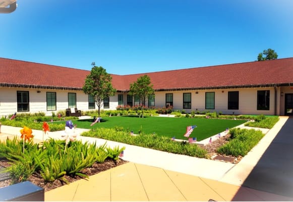 A sunny courtyard with green grass, flowers, and American flags at The Bradford Memory Care.