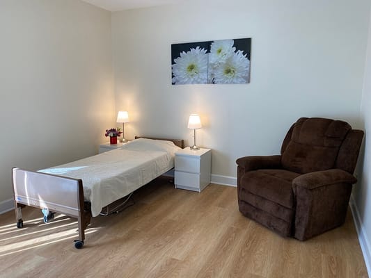 A well-lit room featuring a bed, side table with lamp, and armchair