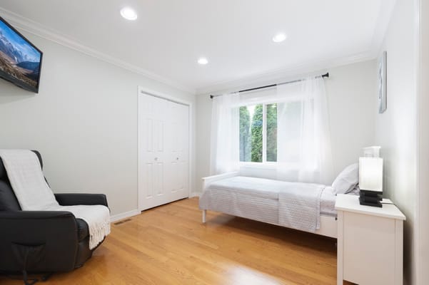 A cozy residential room with a bed and a chair, featuring natural light from a window.