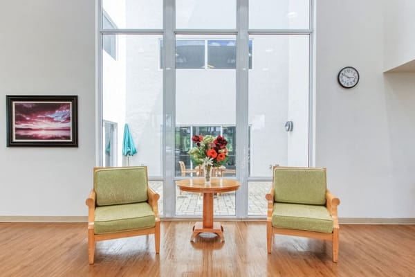 Two green chairs and a small table in a bright lobby area with large windows.