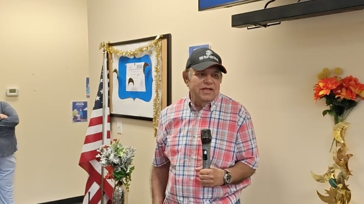 A man speaking at a community event with an American flag and decorations in the background