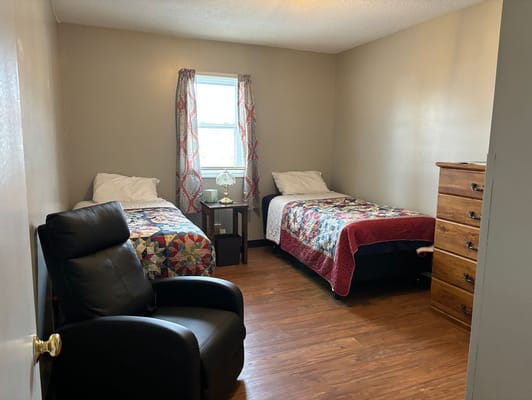 A shared bedroom with two beds and a chair in a senior living facility.
