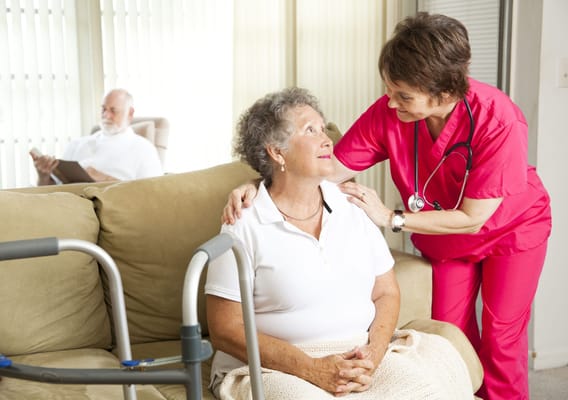 A nurse comforts an elderly woman in a cozy living room.