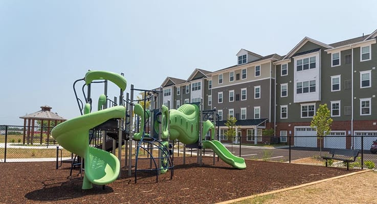 Green playground equipment in a fenced area near apartment buildings.