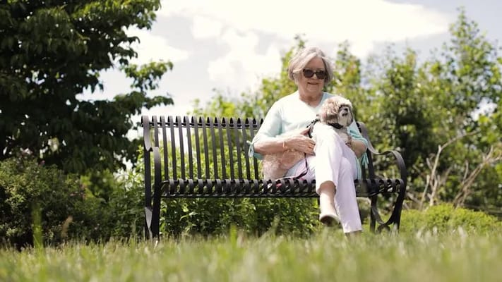 An elderly woman sitting on a bench with a dog in a garden