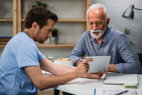 A caregiver showing a tablet to an elderly man at a table with snacks.