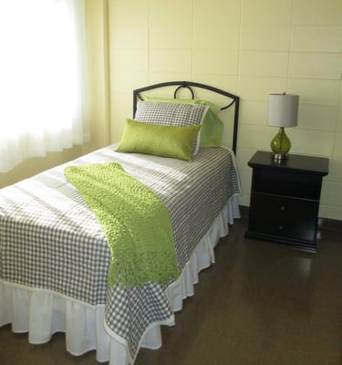 A neatly made single bed with a green throw blanket and a lamp on a nightstand.
