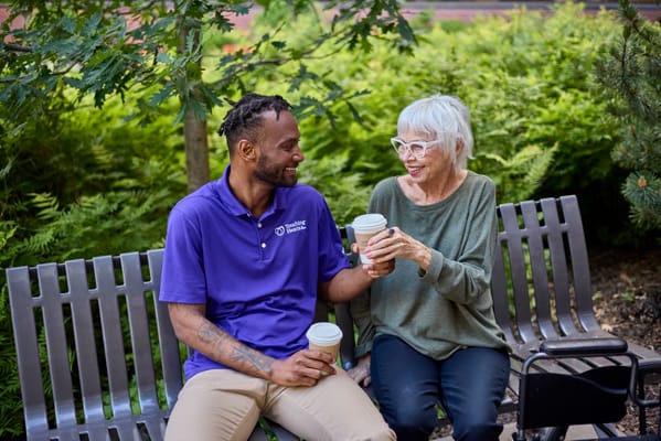 A caregiver and a senior woman sharing coffee on a bench in a garden setting.