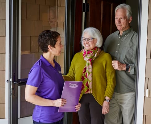 A staff member greets a senior couple at the entrance of a home.
