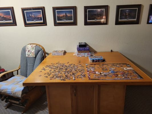 A wooden table with a jigsaw puzzle partially completed, surrounded by rocking chairs and framed pictures