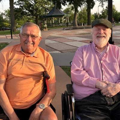 Two seniors smiling in wheelchairs at a park