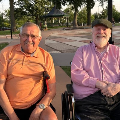 Two seniors smiling in wheelchairs at a park