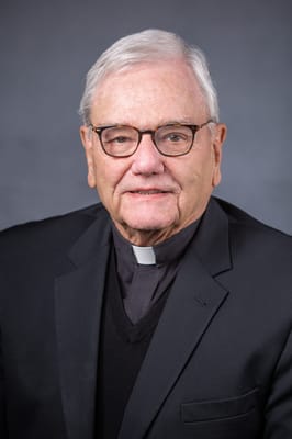 A senior man in eyeglasses wearing a clergy suit