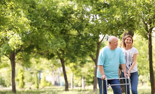 Senior man using a walker in a park with staff assistance