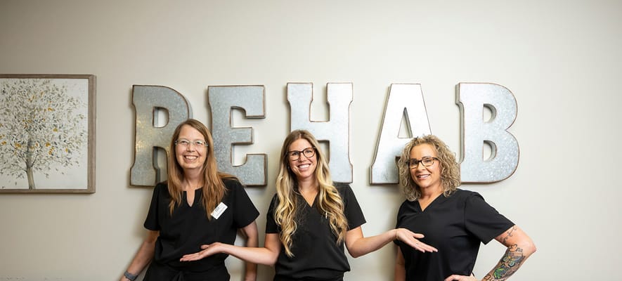 Three staff members posing in front of a 'REHAB' sign