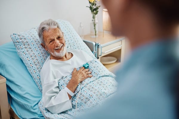 A smiling elderly man in a hospital bed with a caregiver in view