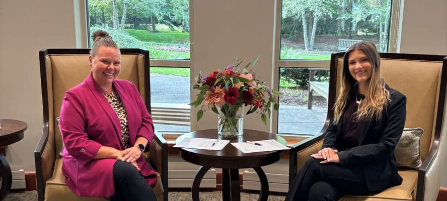 Two staff members smiling in a common area with floral arrangement