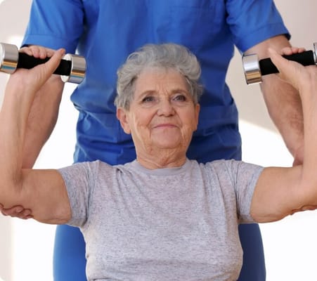An elderly woman exercising with weights, assisted by a caregiver