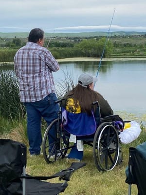 Two people fishing by a pond, one in a wheelchair