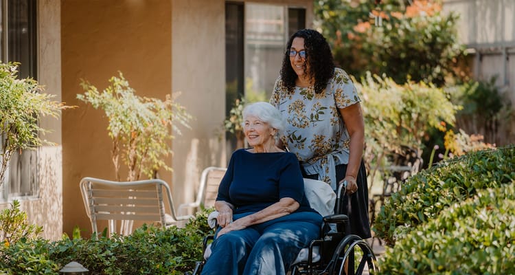 A resident smiling in a wheelchair being assisted outdoors