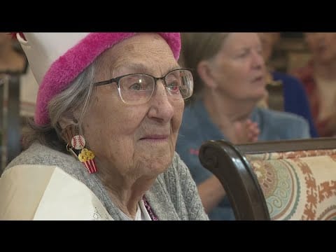 A senior woman with glasses and a pink hat in a social setting