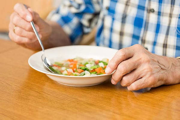 An elderly person's hand reaching for a bowl of vegetable soup
