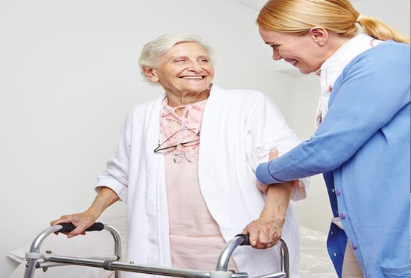 A smiling elderly woman with a walker assisted by a caregiver