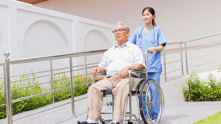 Senior resident being assisted in a wheelchair by a caregiver outdoors