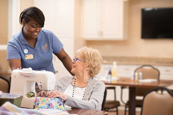 A staff member assists a resident with sewing in an activity room