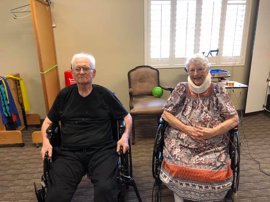 Two smiling residents in wheelchairs in a common area