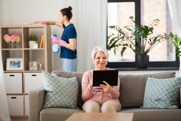Resident using a tablet in a cozy living area with staff cleaning