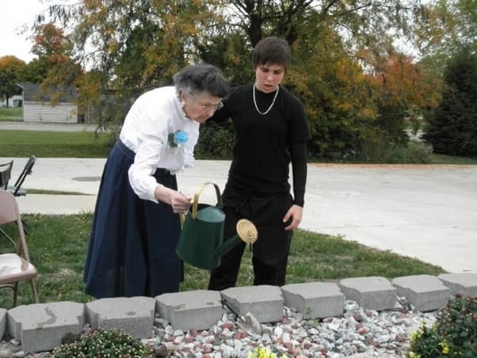An elderly woman watering plants with a young man in a garden