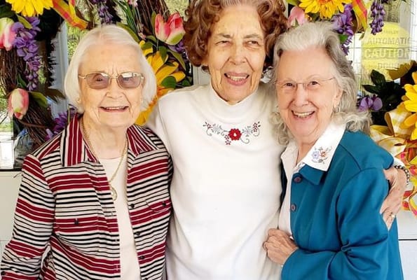Three elderly women smiling together in front of flowers