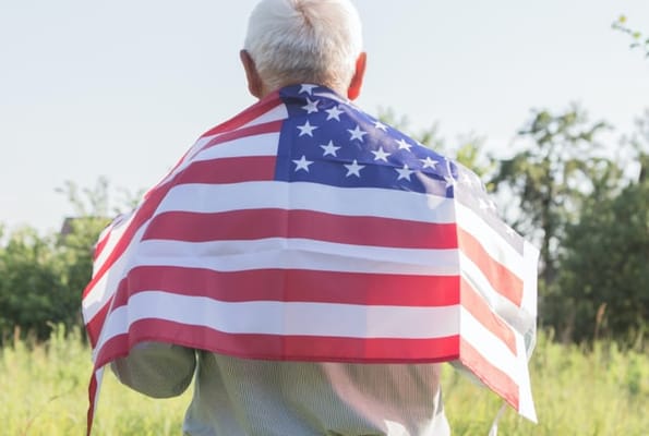 Elderly person wearing an American flag as a cape in a field