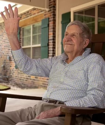 An elderly man waving while seated on a porch