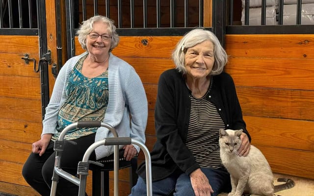 Two residents smiling with a cat in a barn setting