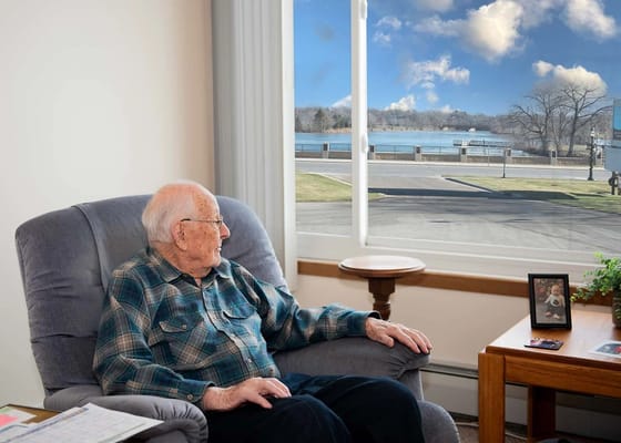 An elderly man looking out a window at a lake view