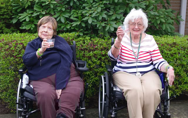 Two elderly residents in wheelchairs raising glasses outdoors