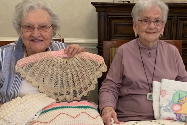 Two residents showing handmade crochet items in an activity room
