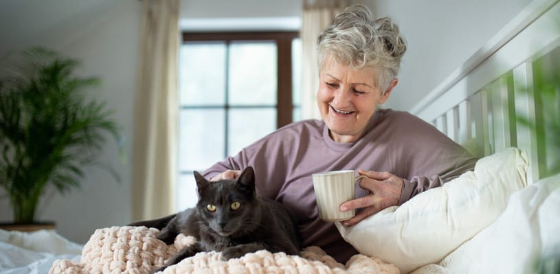 A senior woman sitting on a bed with a cat and a cup