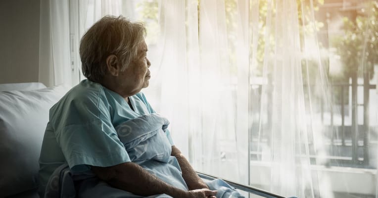 An elderly woman sitting by a window wrapped in a blanket