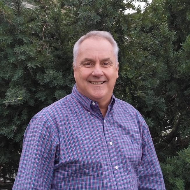 Smiling man standing outdoors near greenery.