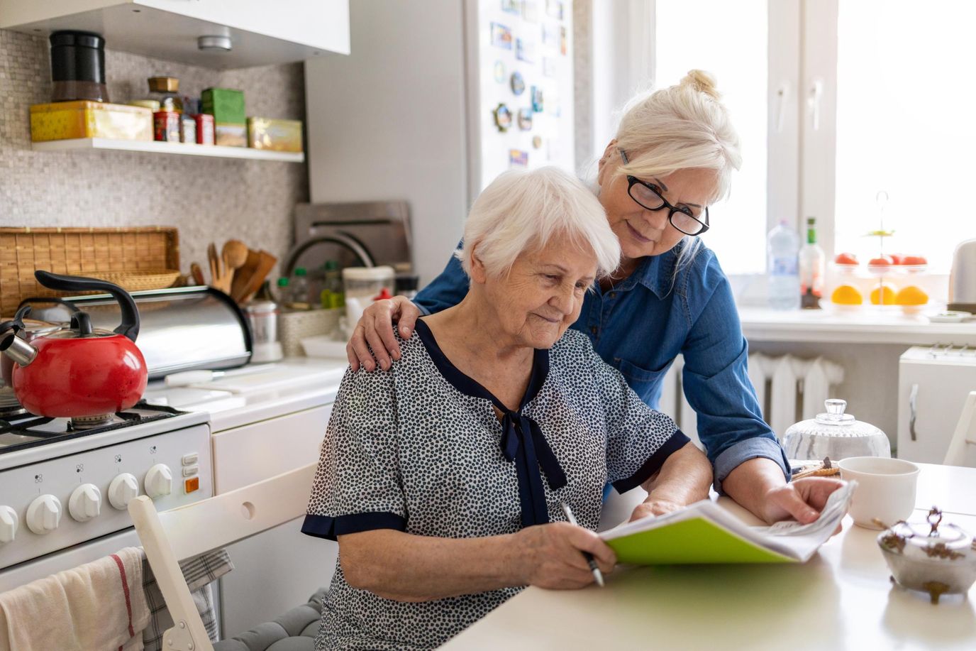 Elderly woman and caregiver reading together in the kitchen.