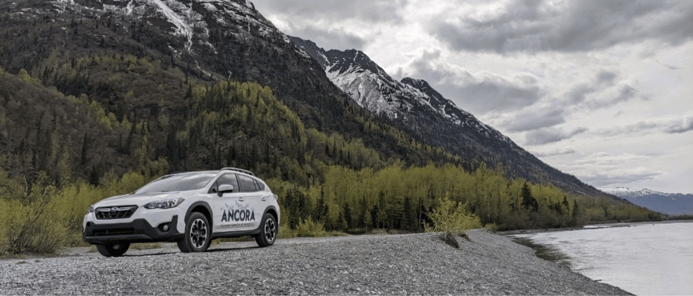 Vehicle parked by a river with mountains in the background.