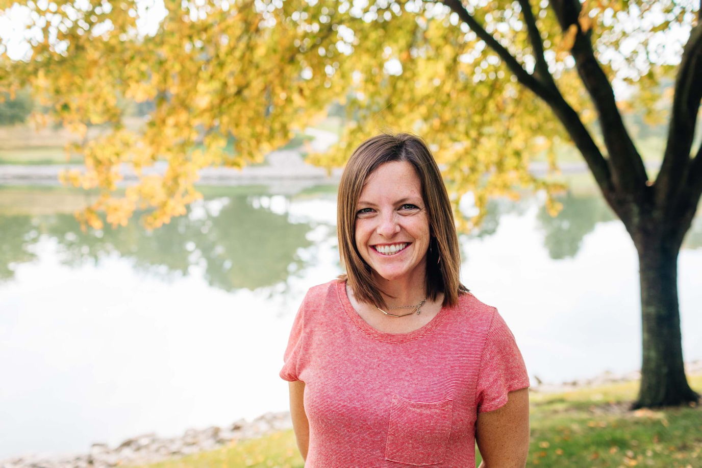 A smiling woman standing by a pond with fall foliage